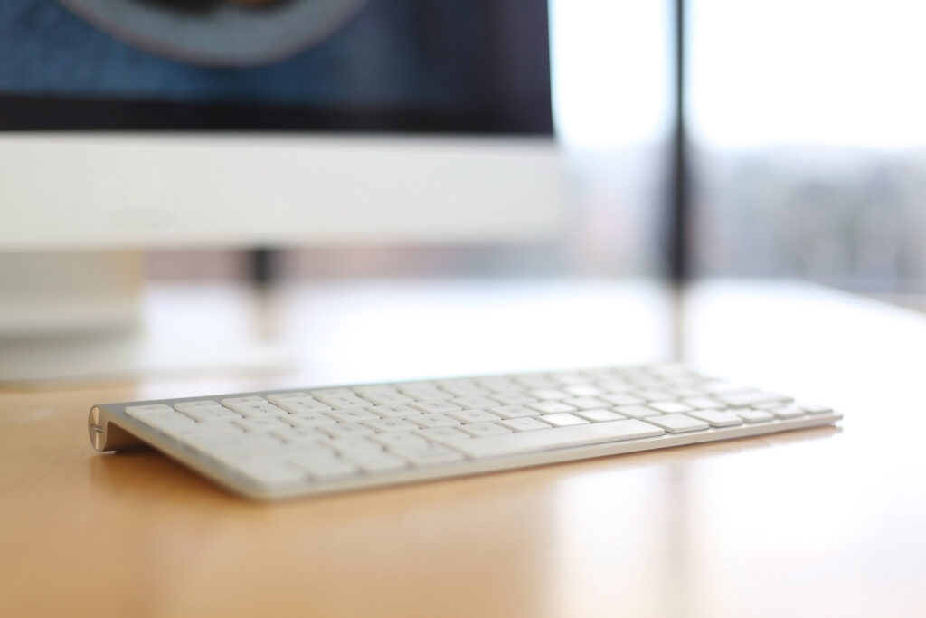 White computer keyboard on a wooden desk, with a blurred monitor in the background, symbolizing digital marketing and technology solutions.